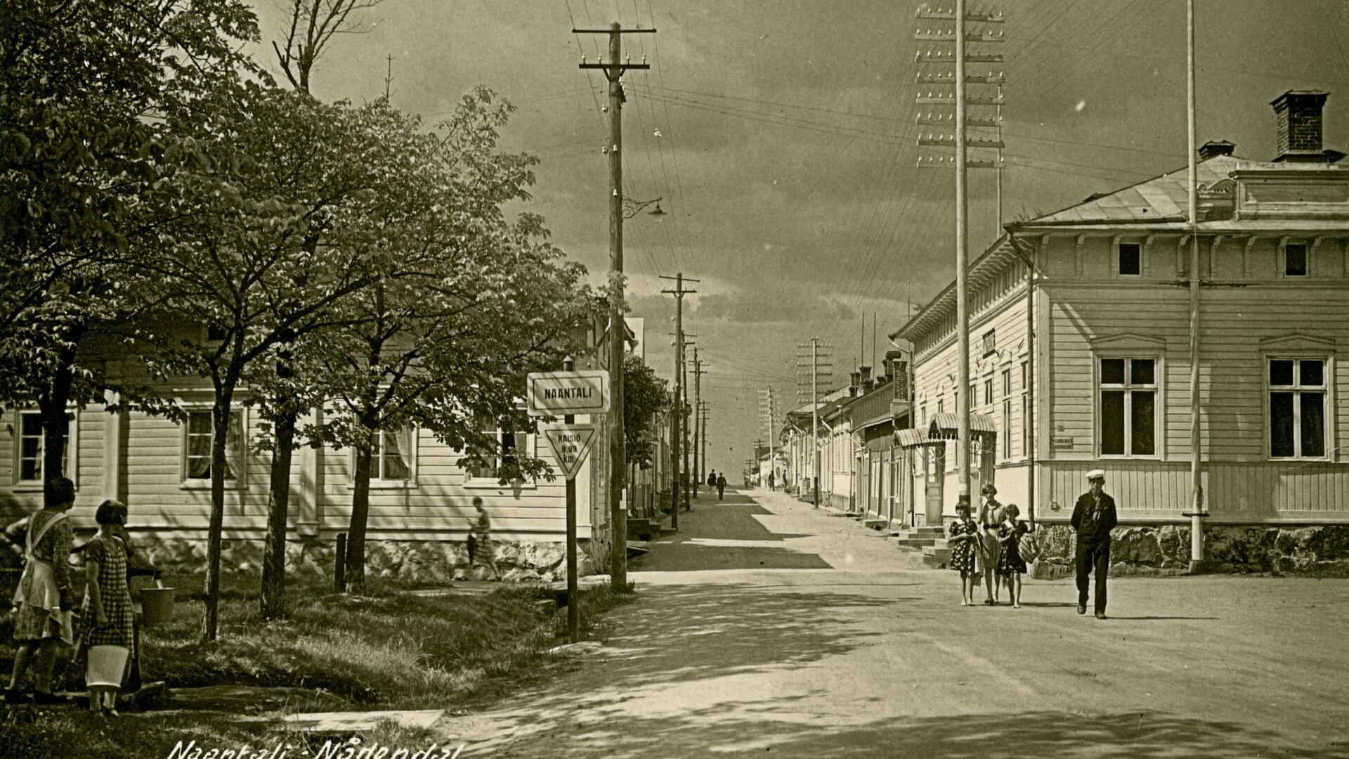 Mustavalkokuvassa katumaisema, jossa puutaloja ja vasemmalla puita ja kaksi tyttöä pumppaamassa vettä kaivosta