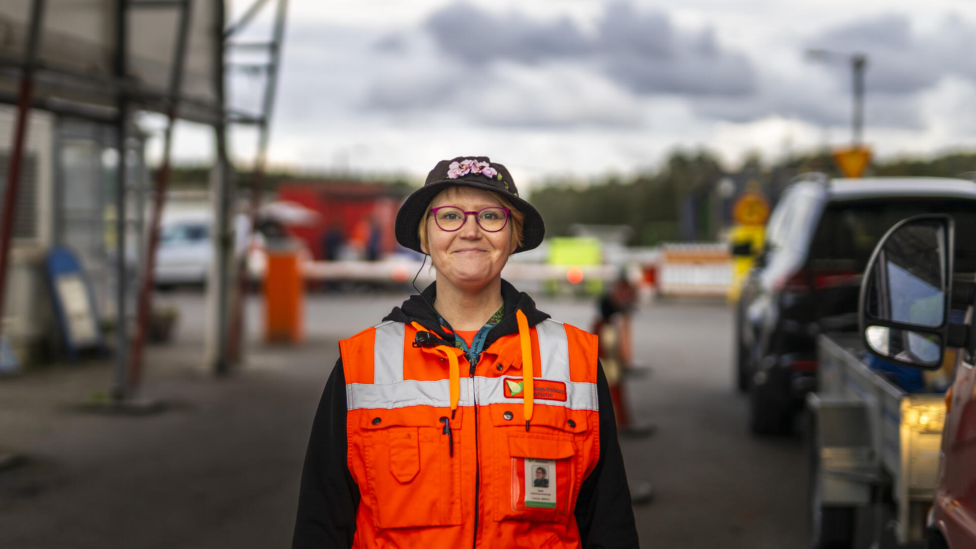 Jätehuollon työntekijä hymyilee kameralle huomioliivi päällä. Taustalla jäteasema.