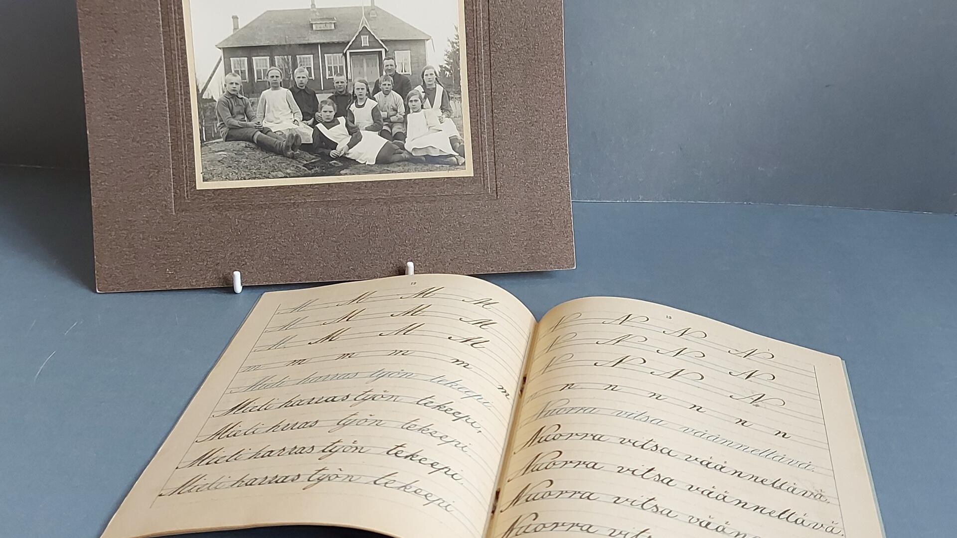 siniharmaalla taustalla avoin kouluvihko ja sen takana mustavalkoinen luokkakuva