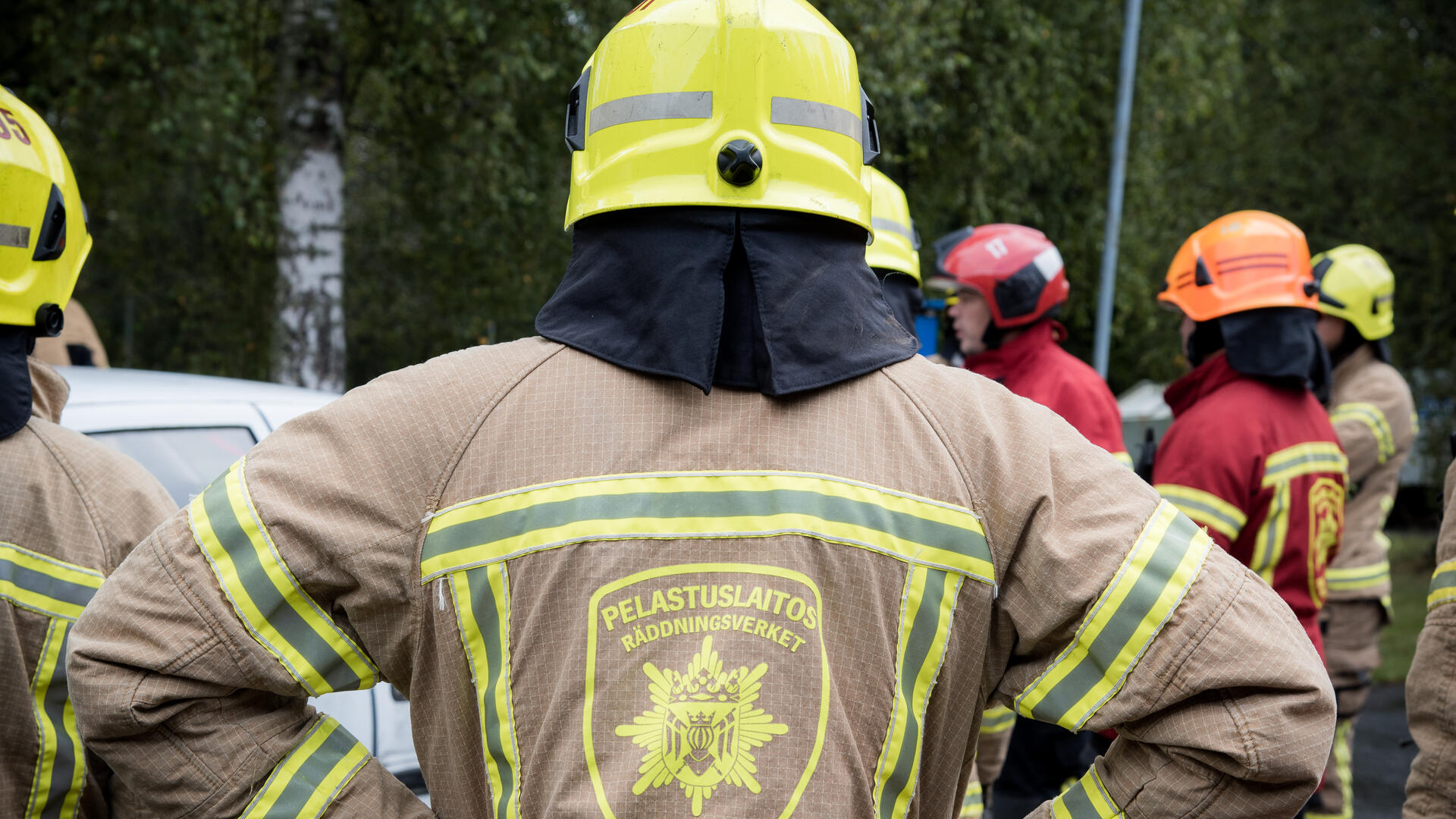 Palomies seisoo selin kameraan, hänen edustallaan lisää palomiehiä.