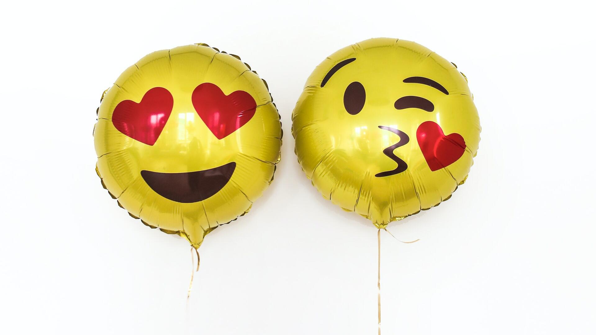 Kaksi keltaista emoji-vappupalloa - toisen silmät ovat sydämet, toinen pussaa.
