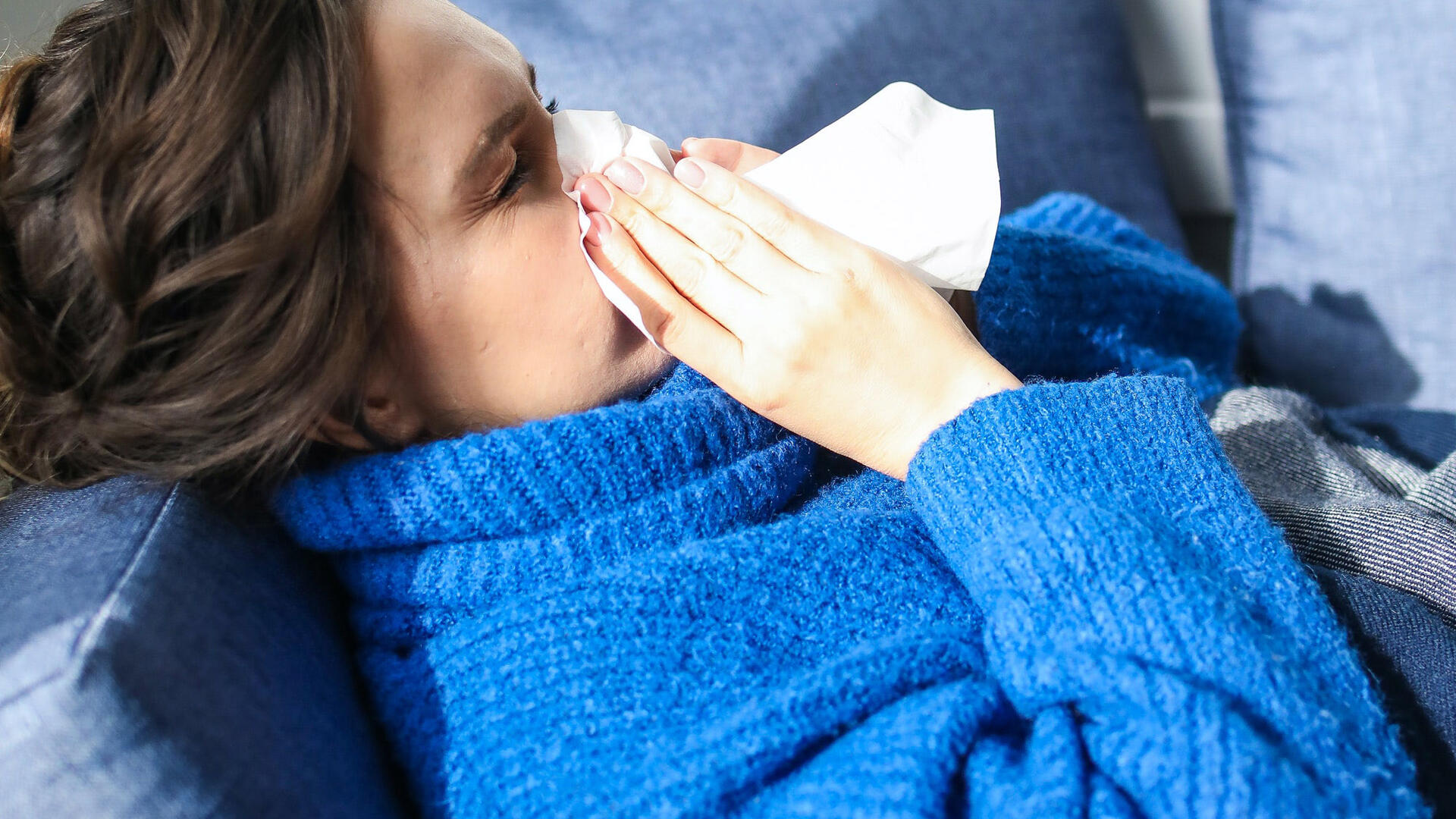 Nainen niistää nenää ja sairastaa flunssaa kotisohvalla.