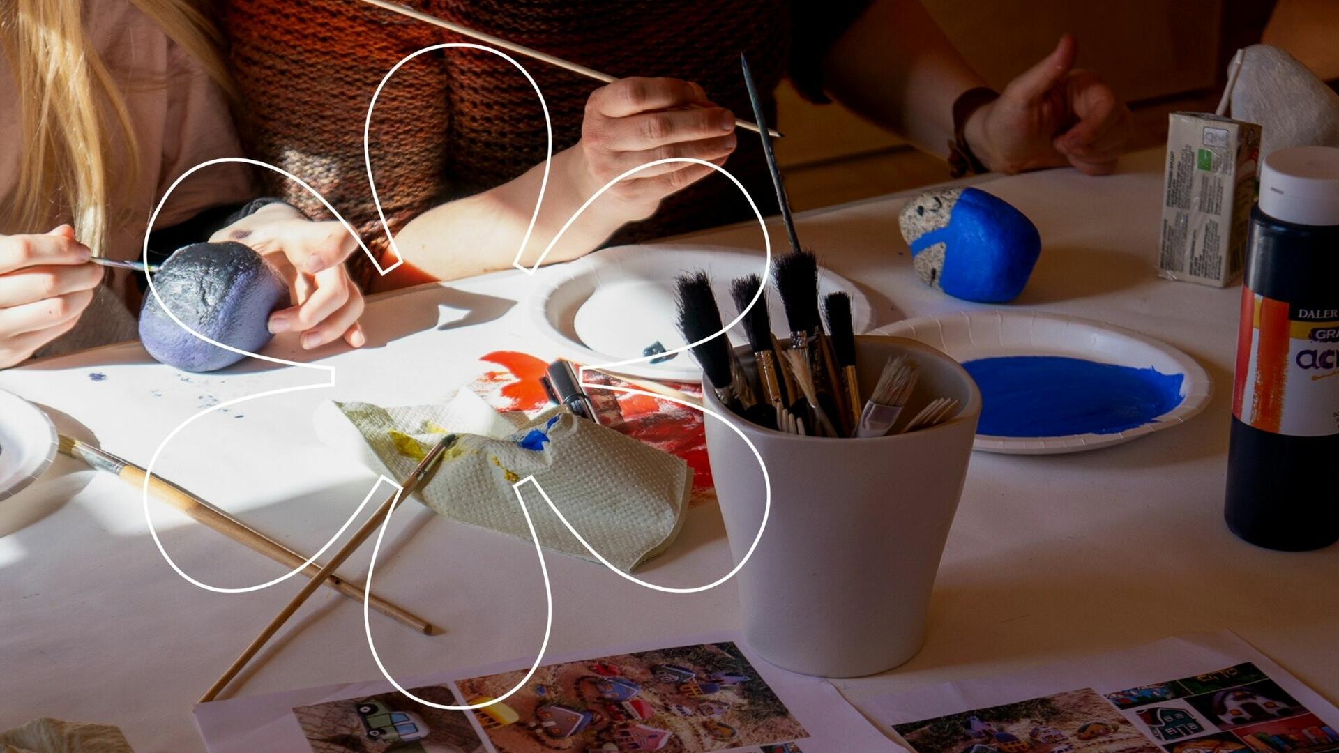 Lapsi ja aikuinen istuvat pyödän ääressä maalaamassa kiviä, pöydällä on kupissa useita pensseleitä, isoja maalituubeja ja papereita, joissa on maalattujen kivien kuvia.