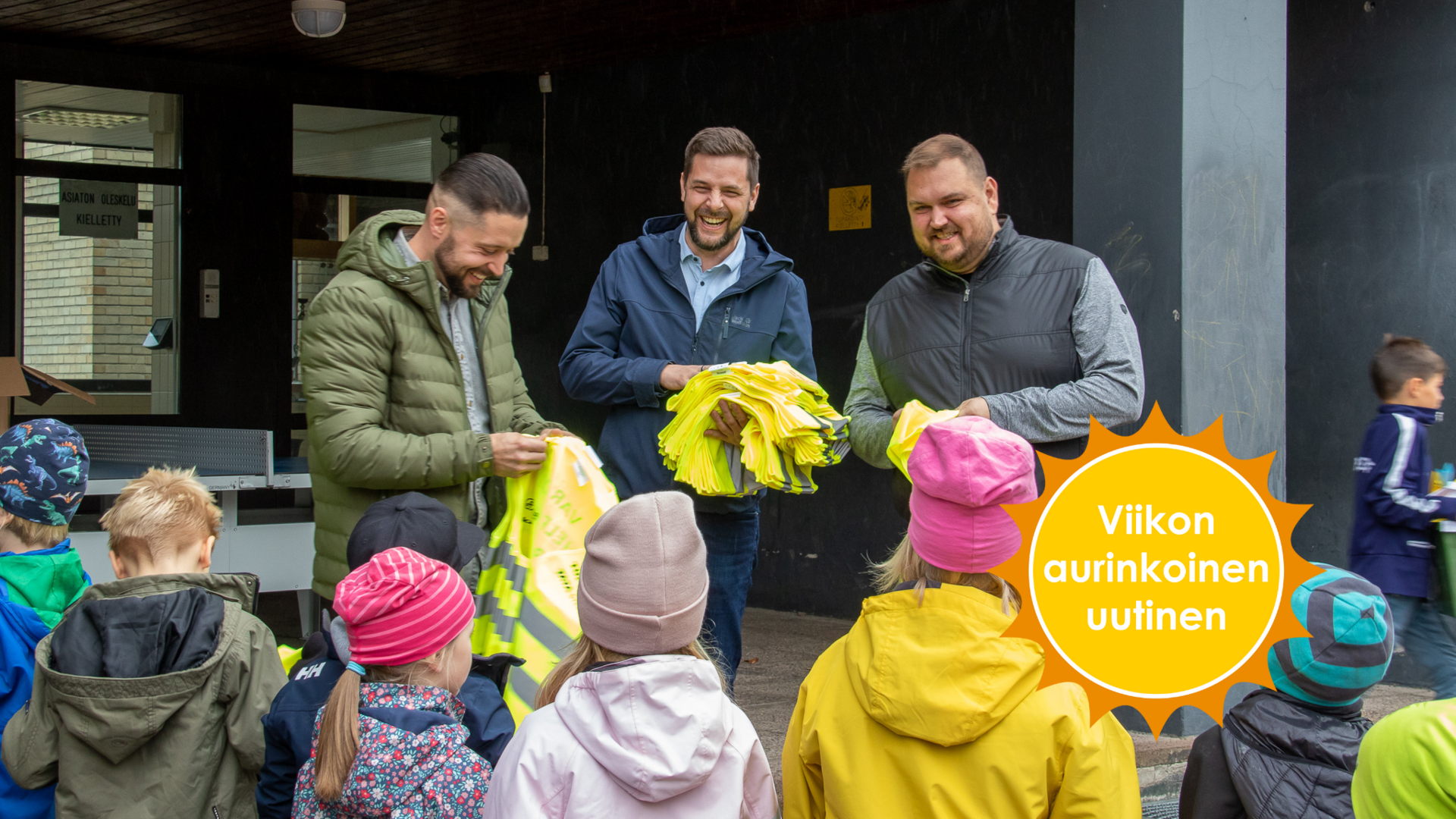 Kolme iloista miestä seisoo lapsiryhmän edessä ja jakaa keltaisia heijastinliivejä.