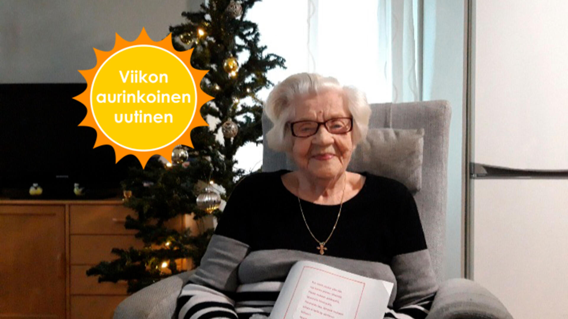 Hymyilevä ikäihminen istuu joulupaketin kanssa joulukuusen edessä. Viikon aurinkoinen uutinen