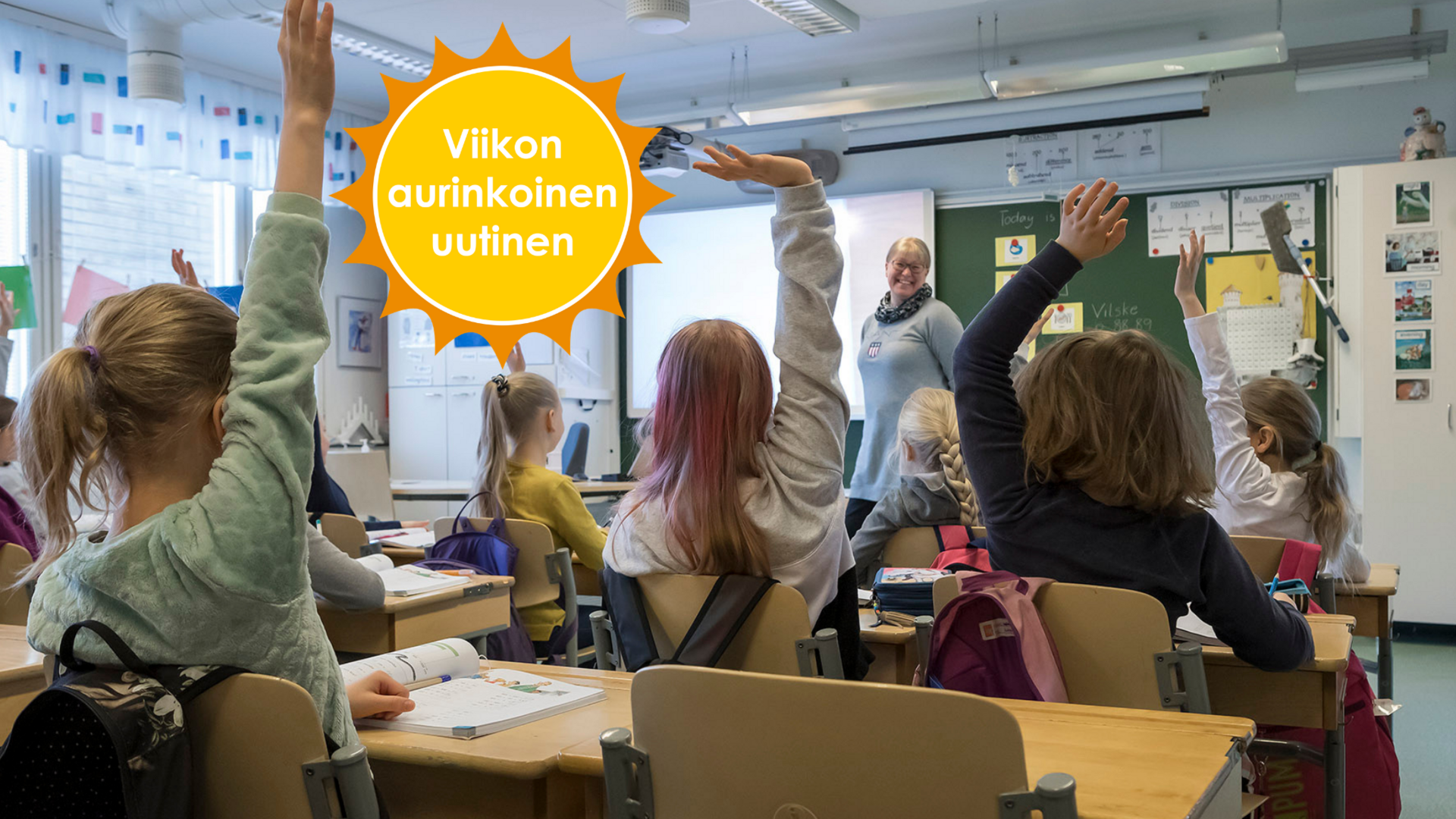 Koululaisia istuu luokassa ja viittaa, opettaja seisoo pulpettien ja taulun välissä ja hymyilee. Viikon aurinkoinen uutinen -logo. 