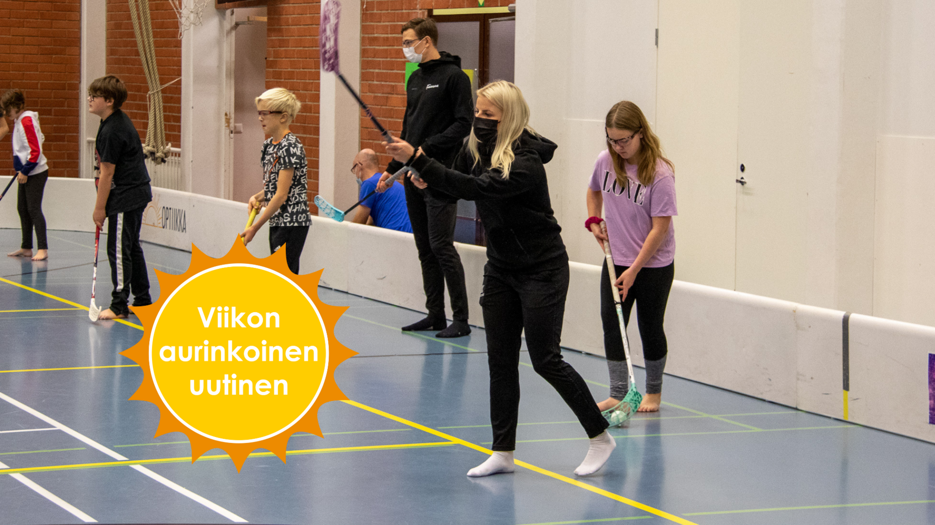 Mustiin pukeutunut Sanni Niemisen maila on koholla pallon laukaisun merkiksi, rivissä hänen kanssaan seisoo lapsia sekä Eemeli salin mailojen kanssa liikuntasalissa. Viikon aurinkoinen uutinen -logo.