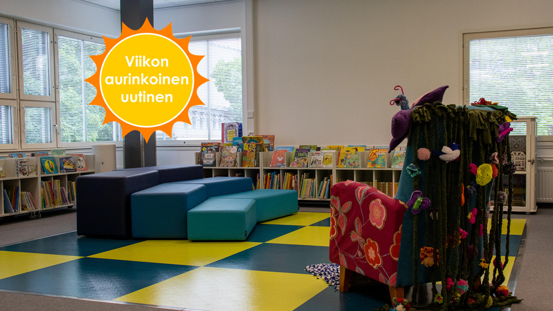Kirjaston lastenosaston sini-keltaruudullinen lattia, jonka kulmassa on monivärinen ja monista kankaista ja materiaaleista koostettu nojatuoli. Vastapäätä sinisiä istuimia monessa tasossa ja matalia hyllyjä lastenkirjoja. Viikon aurinkoinen uutinen-logo.