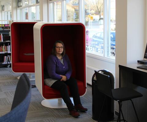 Nainen istuu kirjastossa pehmustetussa kuution muotoisessa tuolissa. 