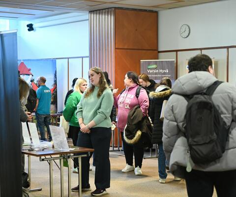 Ihmisiä salissa katselemassa ja juttelemassa messupisteillä.