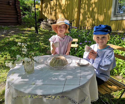 Museon pihalla vietetään lasten museopäivää, jolloin tyttö ja poika vanhan ajan vaatteisiin pukeutuneina juovat kahvia katetun pöydän ääressä.