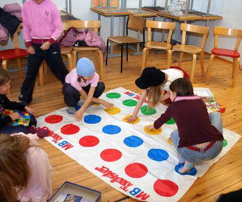 Nuoria lapsia Twister-pelialustan päällä kädet ja jalat ristissä ja eri puolilla värikkäillä palloilla.