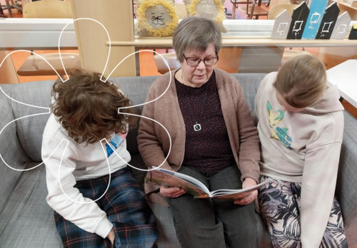 Vapaaehtoinen koulumummo lukee kahdelle oppilaalle kirjaa.