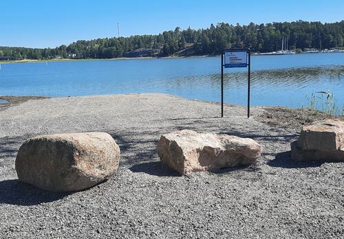Rantaan vie tie, jonka eteen on laitettu isoja kiviä estämään kulku. Kyltti osoittaa koirien uimapaikasta.