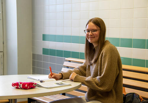 Nuori oppilas istuu pöydän ääressä tehden muistiinpanoja vihkoon, kirja vieressä.