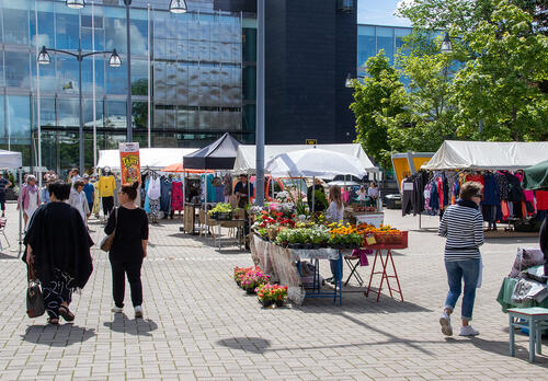 Aurinkoisena kesäpäivänä ihmisiä kulkee torilla, jossa on useita myyntikojuja.