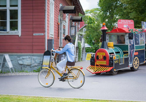 Nuori nainen pyöräilee jopolla Naantalin Vanhassakaupungissa, taustalla kaupunkijuna sekä punainen puutalo.