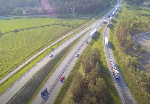 Ylhäältä kuvattuna näkyy E18-tie, jossa rekat ja autot ajavat. Kaistoja liittyy tiehen, ympärillä on vehreää pelto- ja metsämaisemaa.