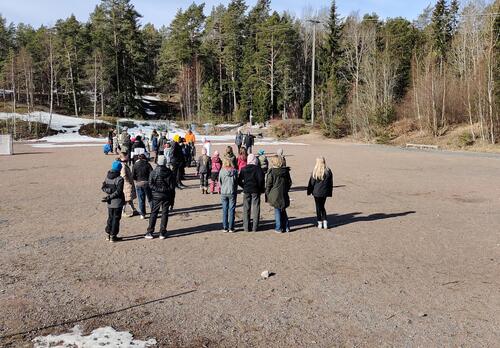Oppilaita seisoo heikkakentällä koulun pihalla kuuntelemassa aikuisia.