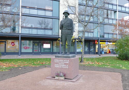 keskellä sotilaspatsas kivipaadella, ympärillä puistomaisemaa ja taustalla kerrostalo, jonka alakerrassa liiketiloja