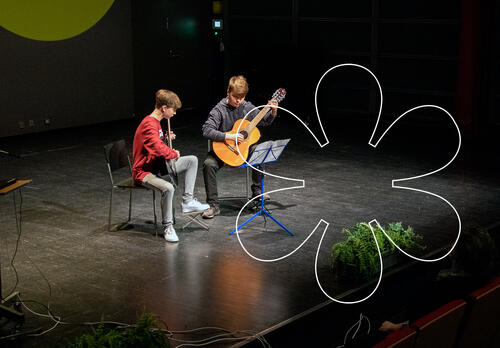 Kaksi nuorta poikaa istuu vierekkäin valokeilassa lavalla kitaraa soittaen, edessään nuottiteline. Kuvan päällä Onnellinen aurinko -elementti.