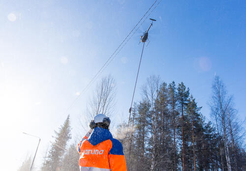 Carunan työntekijä seisoo työmaakypärä, kuulosuojaimet ja oranssi takki päällä metsän edustalla, sähkölinjojen yläpuolella on helikopteri.