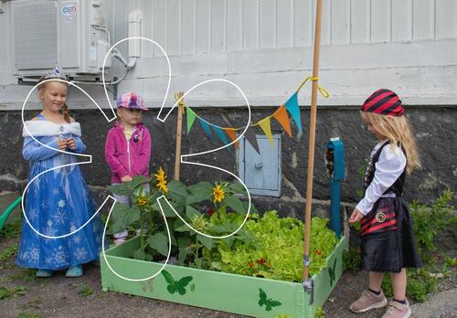 Kolme lasta istutuslavan ympärille kerääntyneenä, laatikossa on pieni kasvimaa ja se on koristeltu mm. värikkäin viirein.