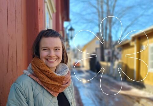 Hymyilevä Heidi Häyry Naantalin Vanhankaupungin puutalojen keskellä, kuvan päällä Onnellinen aurinko -elementti.