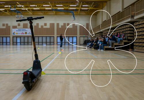 Sähköpotkulauta Maijamäen liikuntahallissa, etäänpänä Sami Kivilä puhumassa nuorelle yleisölle. Kuvassa Onnellinen aurinko -elementti.