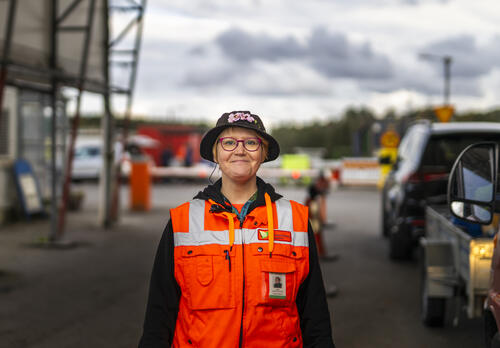 Jätehuollon työntekijä hymyilee kameralle huomioliivi päällä. Taustalla jäteasema.