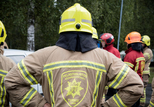 Palomies seisoo selin kameraan, hänen edustallaan lisää palomiehiä.
