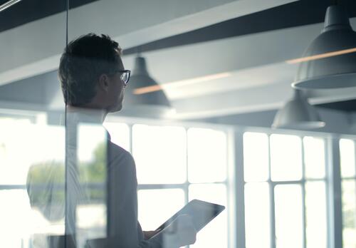 Silmälasipäinen mies seisoo tabletin kanssa lasiseinän takana.