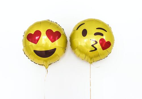Kaksi keltaista emoji-vappupalloa - toisen silmät ovat sydämet, toinen pussaa.