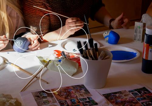 Lapsi ja aikuinen istuvat pyödän ääressä maalaamassa kiviä, pöydällä on kupissa useita pensseleitä, isoja maalituubeja ja papereita, joissa on maalattujen kivien kuvia.