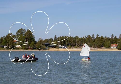 Pieni purjejolla merellä kyydissään kaksi koululaista, lähellä on vene, jossa on enemmän koululaisia ja ohjaaja. Kuvan päällä Onnellinen aurinko -elementti.