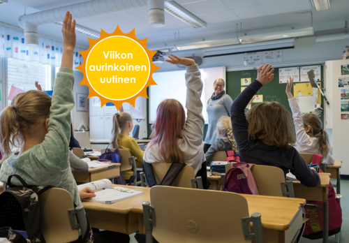 Koululaisia istuu luokassa ja viittaa, opettaja seisoo pulpettien ja taulun välissä ja hymyilee. Viikon aurinkoinen uutinen -logo. 