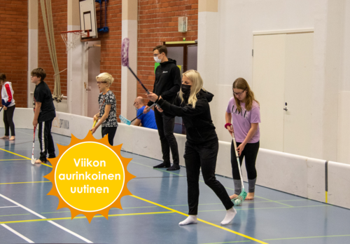 Mustiin pukeutunut Sanni Niemisen maila on koholla pallon laukaisun merkiksi, rivissä hänen kanssaan seisoo lapsia sekä Eemeli salin mailojen kanssa liikuntasalissa. Viikon aurinkoinen uutinen -logo.