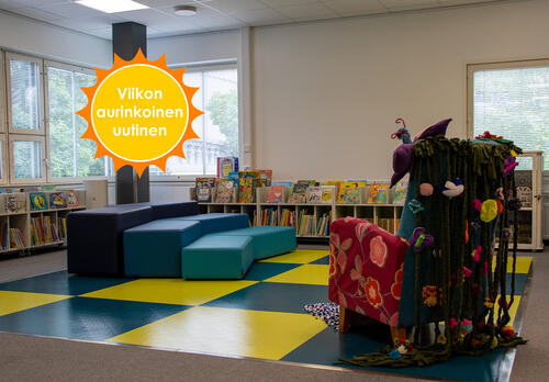 Kirjaston lastenosaston sini-keltaruudullinen lattia, jonka kulmassa on monivärinen ja monista kankaista ja materiaaleista koostettu nojatuoli. Vastapäätä sinisiä istuimia monessa tasossa ja matalia hyllyjä lastenkirjoja. Viikon aurinkoinen uutinen-logo.