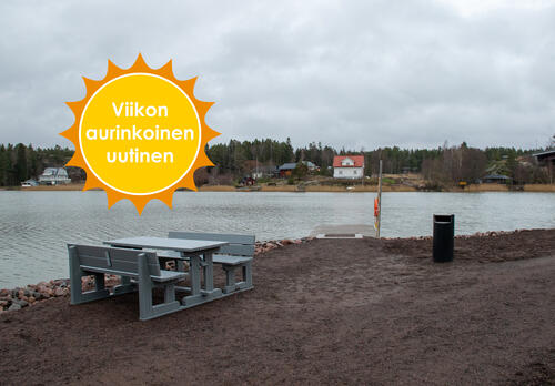 Merimaskun rannassa on harmaa pöytä-penkkiryhmä, roska-astia sekä kalastuslaituri. Viikon aurinkoinen uutinen.