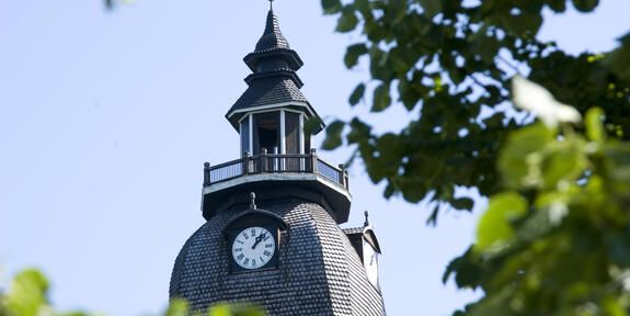 Kirkontorni, jossa kellotaulu, näkyy vihreiden puun lehtien välistä. Taustalla vaaleansininen taivas.