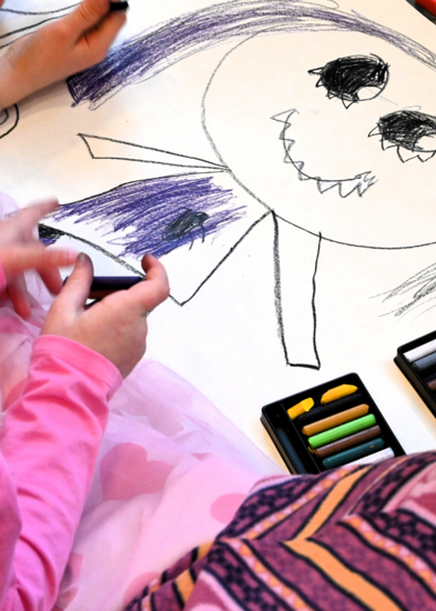 Kaksi oppilasta piirtävät lattialla isoa piirustusta. Kuva otettu olan takaa. Piirustuksessa isosilmäinen hahmo hymyilee.