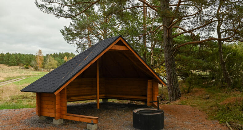 Laavu tulipaikkoineen metsän reunassa, takana peltomaisemaa.