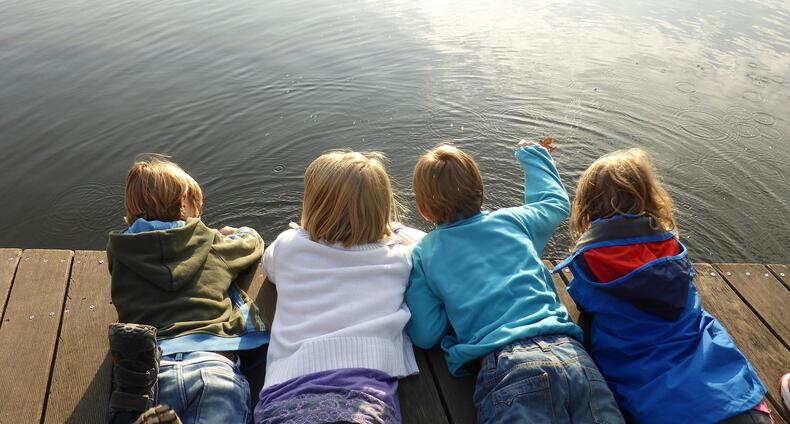 Neljä lasta makoilemassa vatsallaan laiturilla veden äärellä ulkoiluvaatteet päällään.