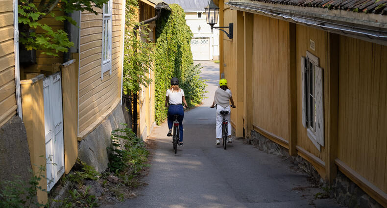 Kaksi nuorta naista pyöräilee Vanhankaupungin puutalojen keskellä.